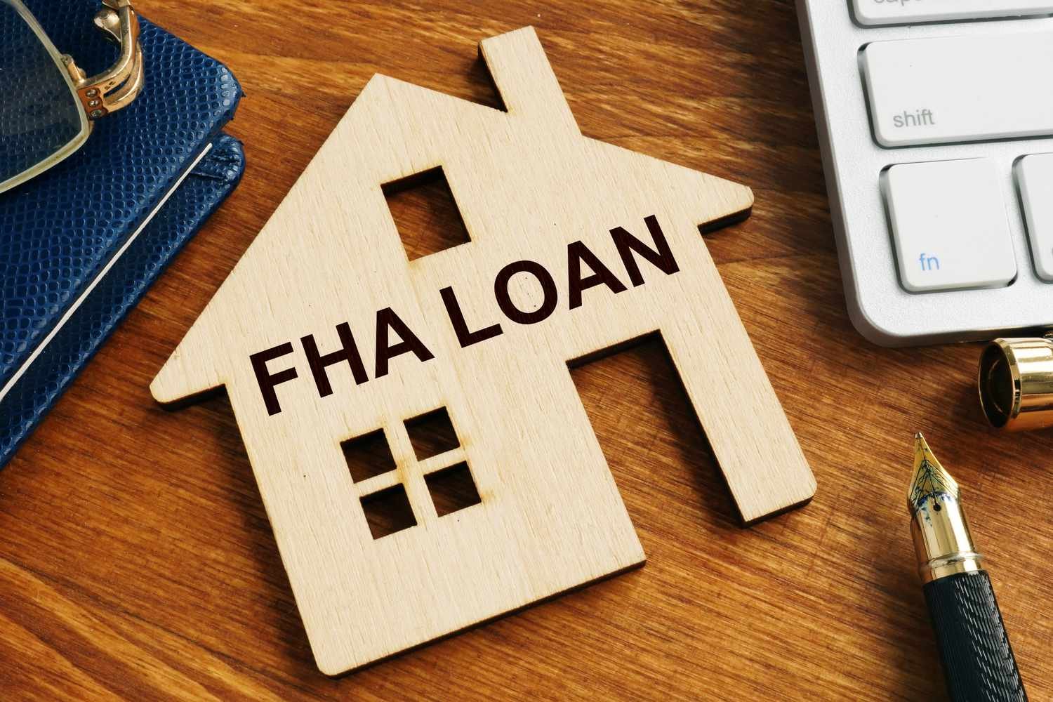 A wooden house cutout labeled "FHA LOAN" on a wooden table, beside a pen, keyboard, and sunglasses.