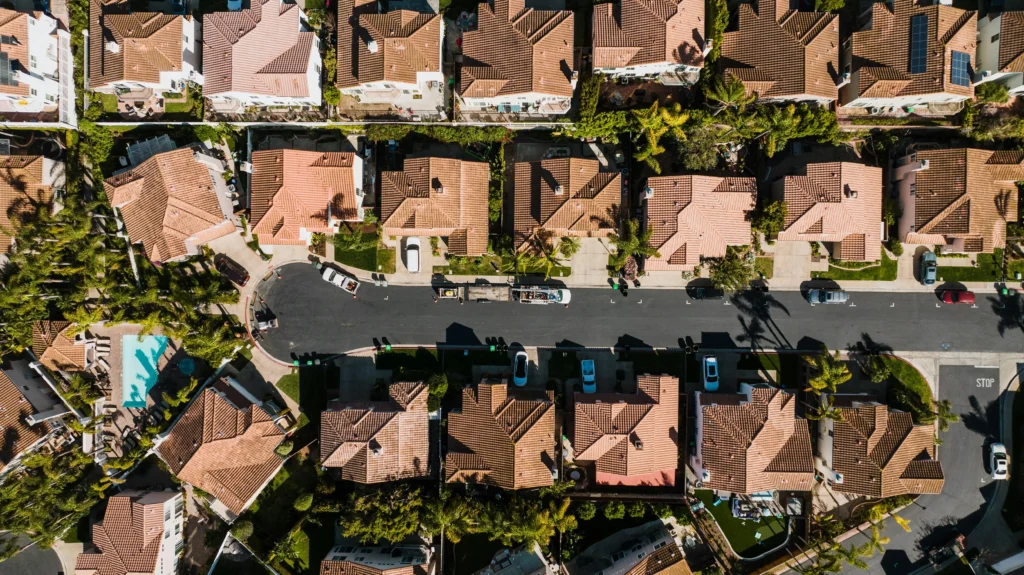 Aerial view of suburban neighborhood with tiled roofs, green lawns, a swimming pool, and cars parked along a winding street.
