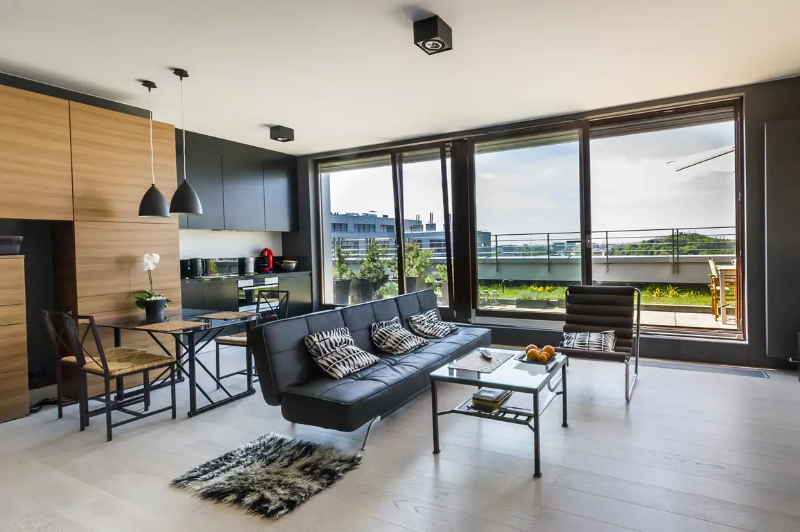 Modern living room with a sleek black sofa, dining area, large windows, and access to a balcony with greenery.