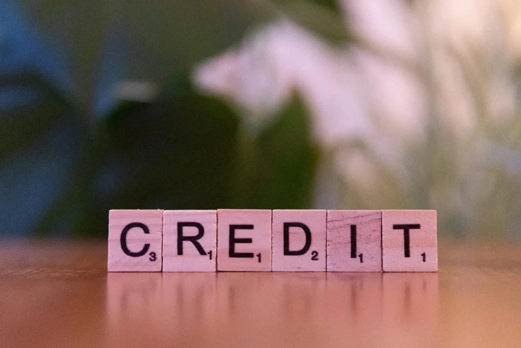 Wooden letter tiles spell out "CREDIT" with visible point values on a wooden surface, set against a blurred green background.