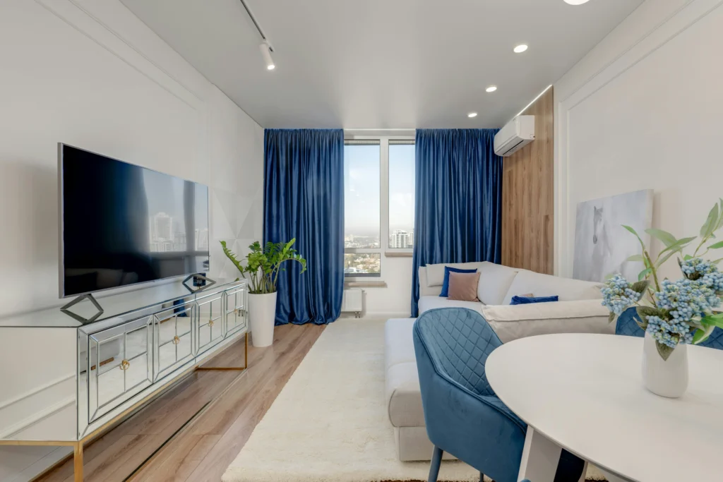 Modern living room with a blue and white color scheme, featuring a TV, plush sofa, and large windows with city views.