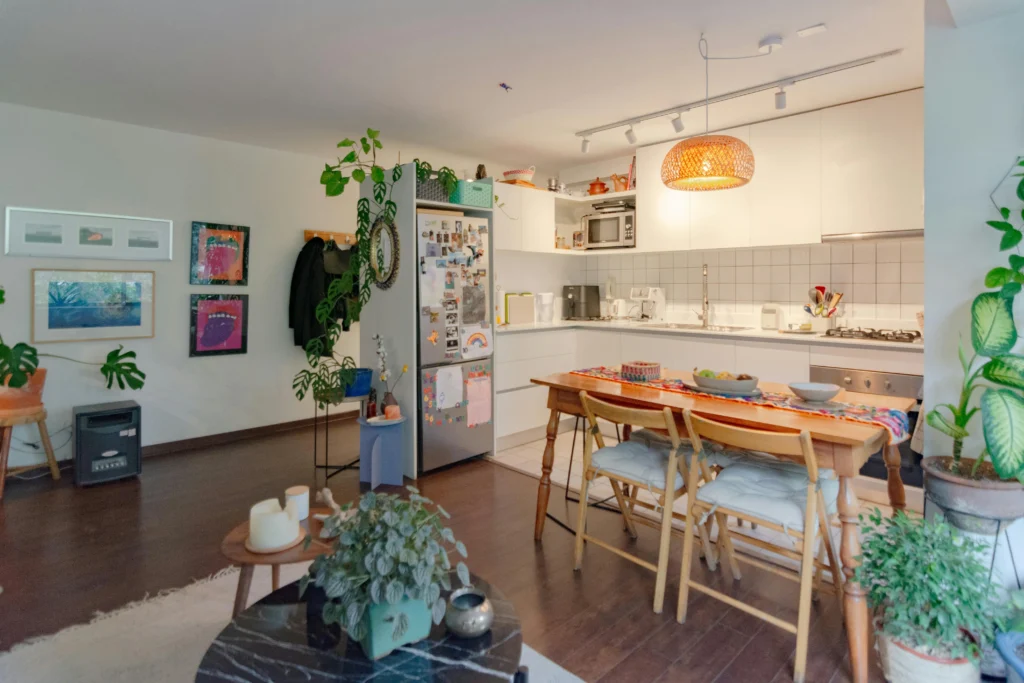 Bright, modern kitchen and dining area with wooden table, colorful decor, plants, and a stylish pendant light. Cozy and inviting atmosphere.