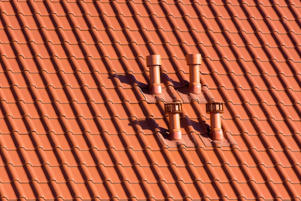 Close-up of a terracotta tiled roof featuring four protruding chimneys, casting shadows on the textured tiles below.