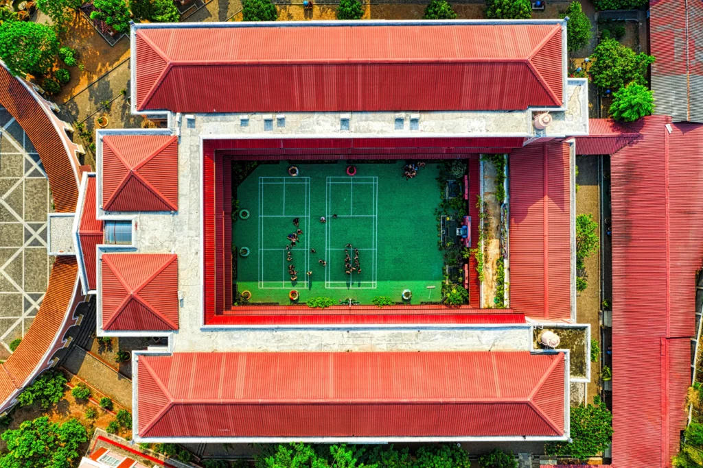 Aerial view of a courtyard surrounded by buildings with red rooftops, featuring tennis courts and students engaged in activities.