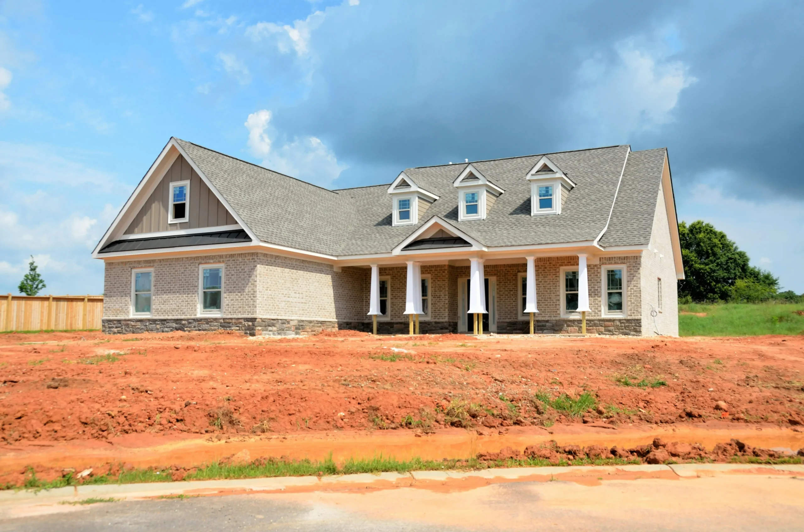 A newly constructed house featuring a mix of brick and stone, with a gray roof and detailed gables, set against a blue sky.