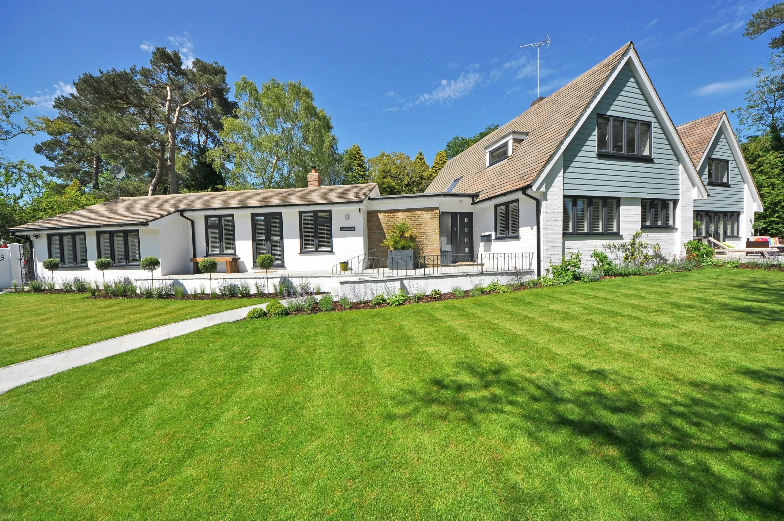 A modern two-story house with a sloped roof, large windows, and a manicured lawn, surrounded by trees and blue sky.