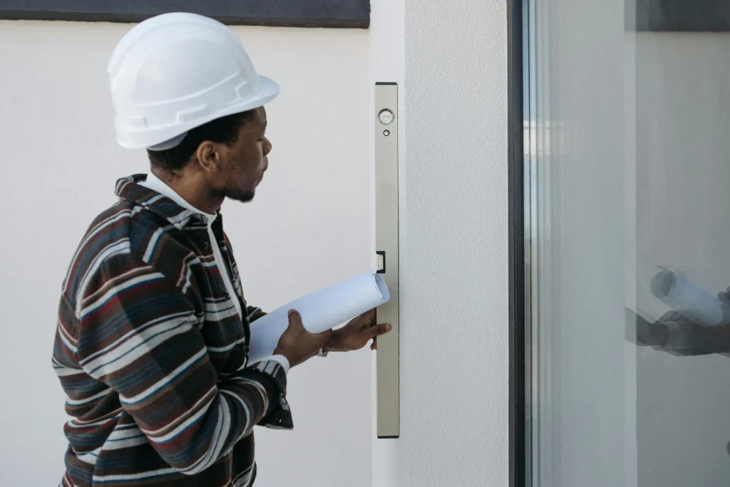 A worker in a hard hat holds architectural plans while checking a wall with a level tool in a modern setting.