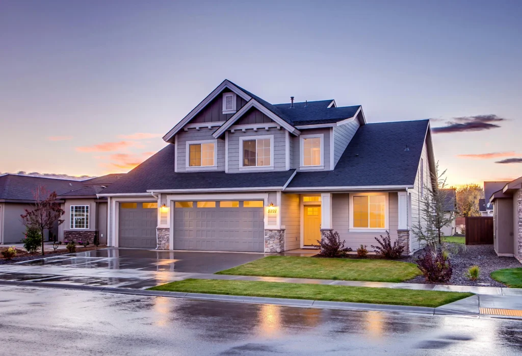 A modern two-story gray house at sunset, featuring large windows, a stone-accented porch, and a reflective driveway.