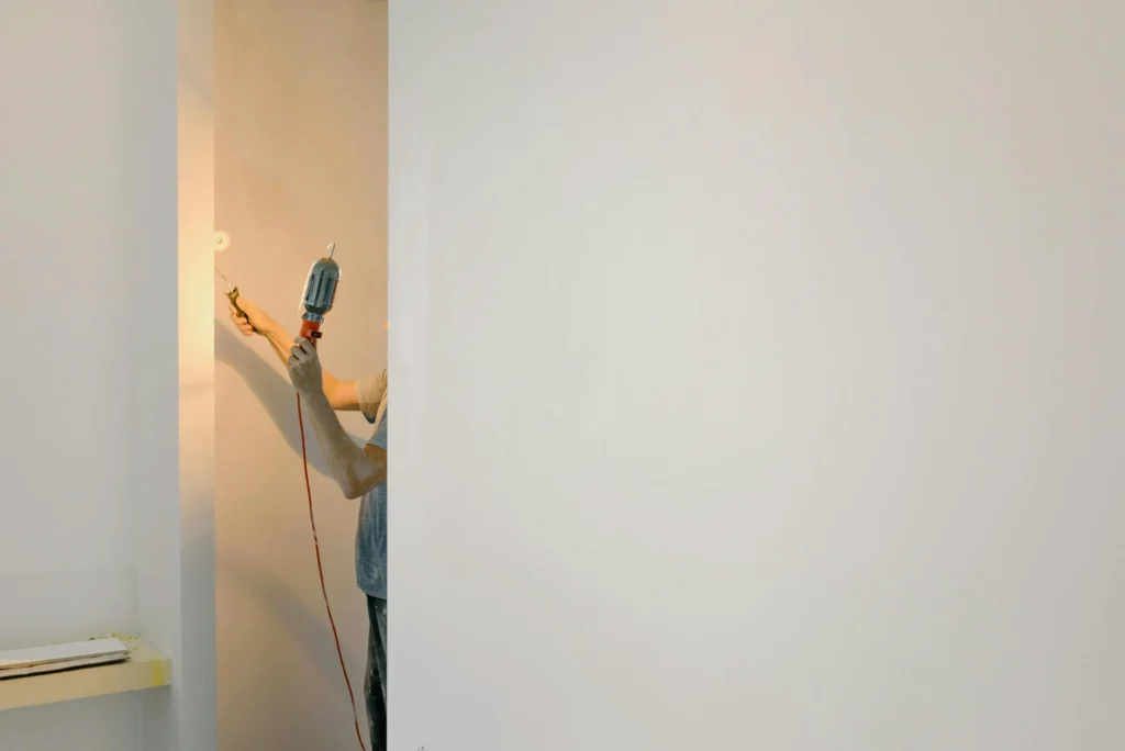 A person holding a paint roller and a drill is painting a wall in a partially completed room with plain white walls.