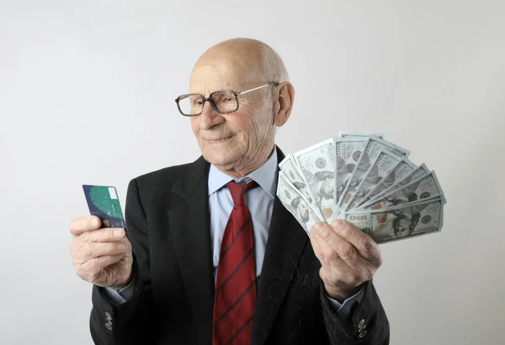 An elderly man in a suit holds a fan of cash in one hand and a credit card in the other, against a plain background.