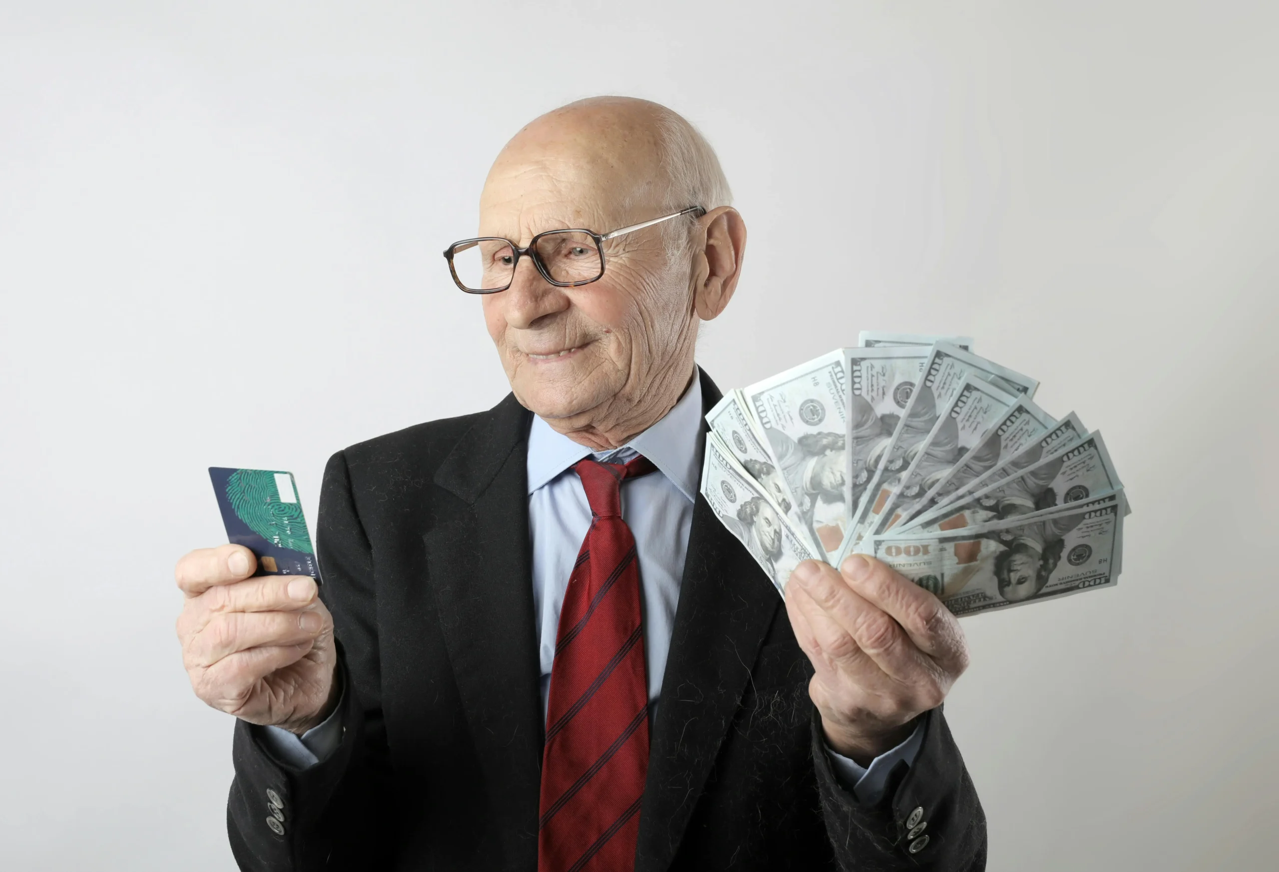 An elderly man in a suit holds a fan of cash in one hand and a credit card in the other, against a plain background.