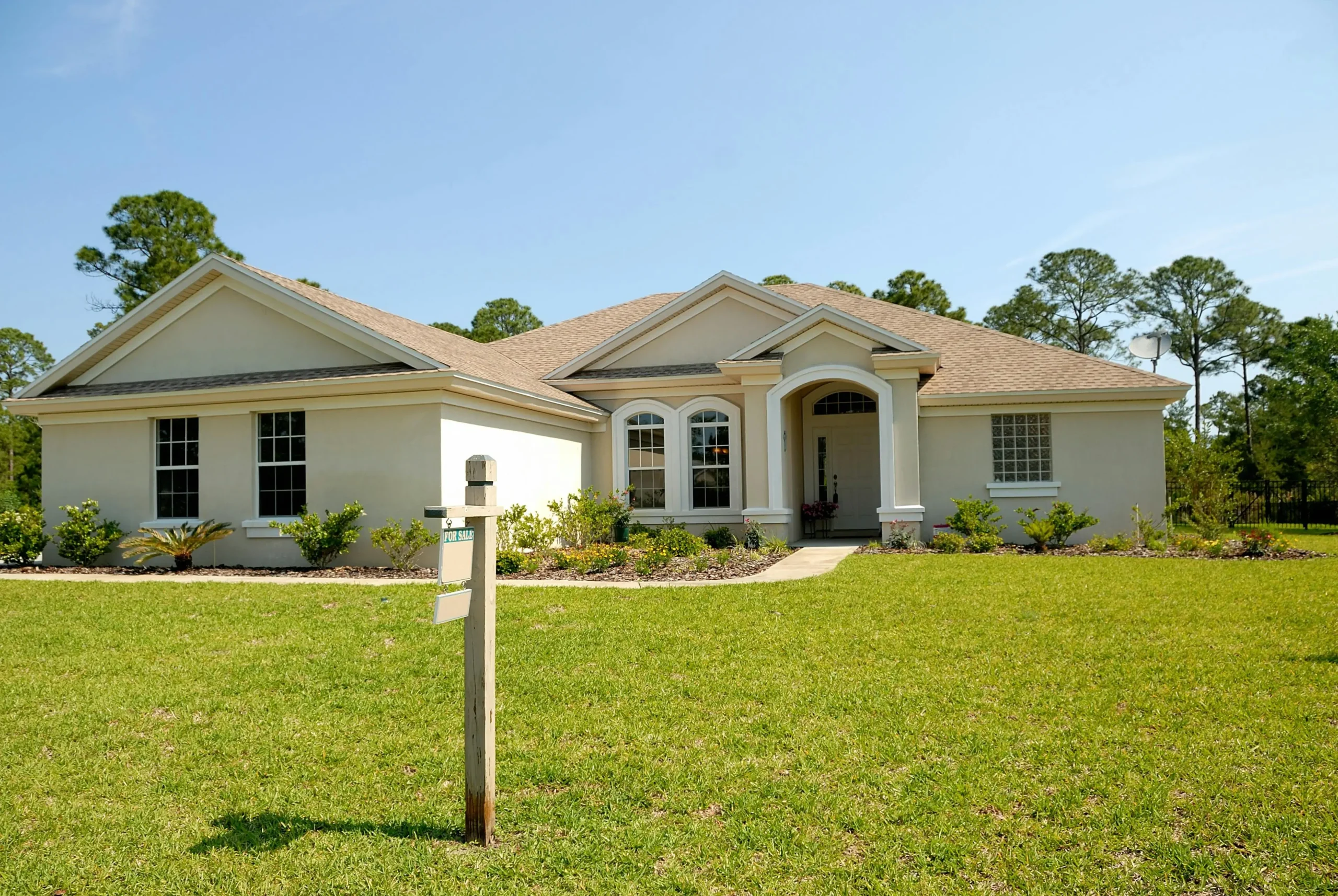 A spacious beige house with a front porch, surrounded by greenery, features a "For Sale" sign in the yard under a clear blue sky.