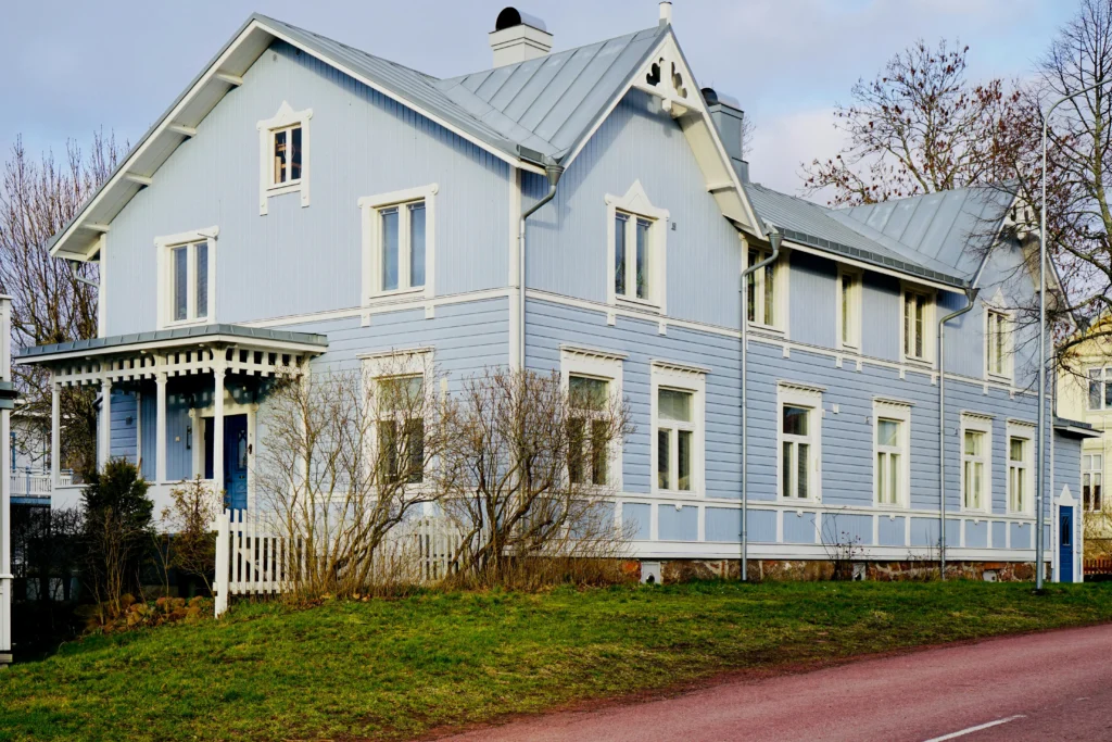 A charming blue wooden house with white trims, featuring a porch and multiple windows, set against a grassy yard.