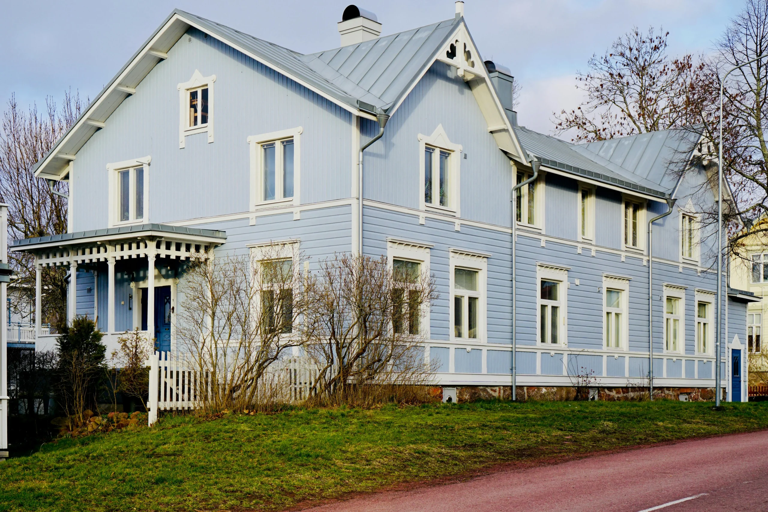 A charming blue wooden house with white trims, featuring a porch and multiple windows, set against a grassy yard.