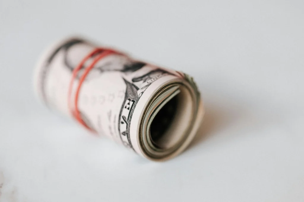 A rolled bundle of cash secured with a red rubber band, resting on a light background.