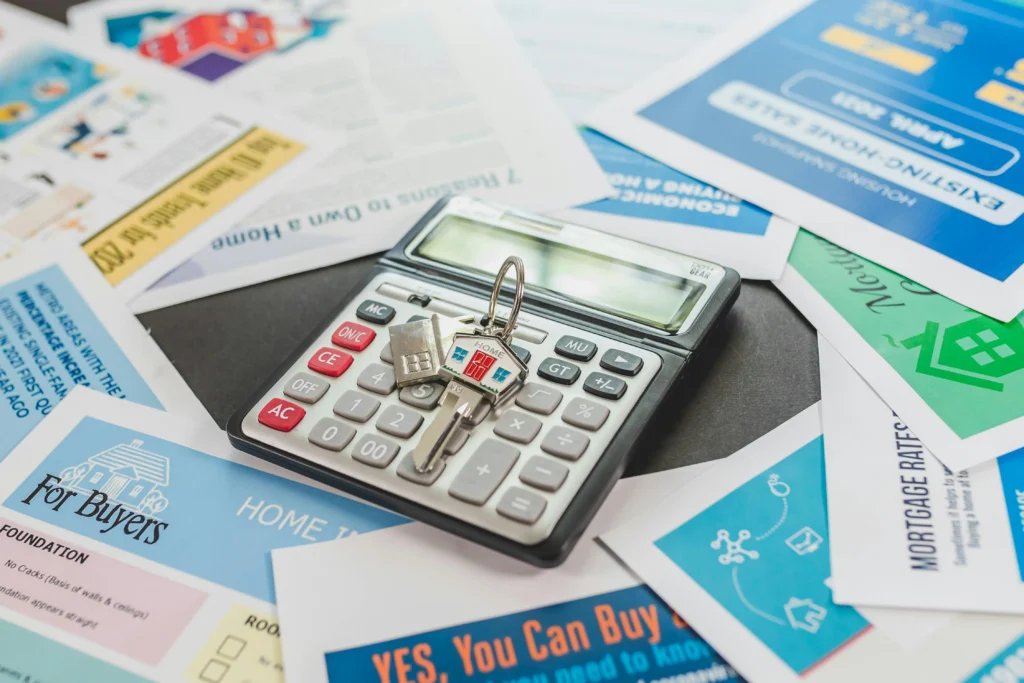 A calculator with a house key rests on a table surrounded by various real estate documents and flyers.