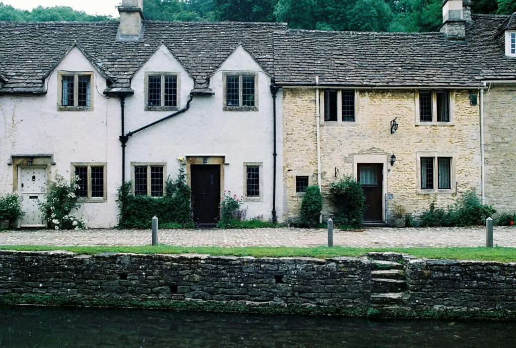 Two quaint cottages with pitched roofs, surrounded by green grass and flowers, beside a stone wall along a gentle riverbank.