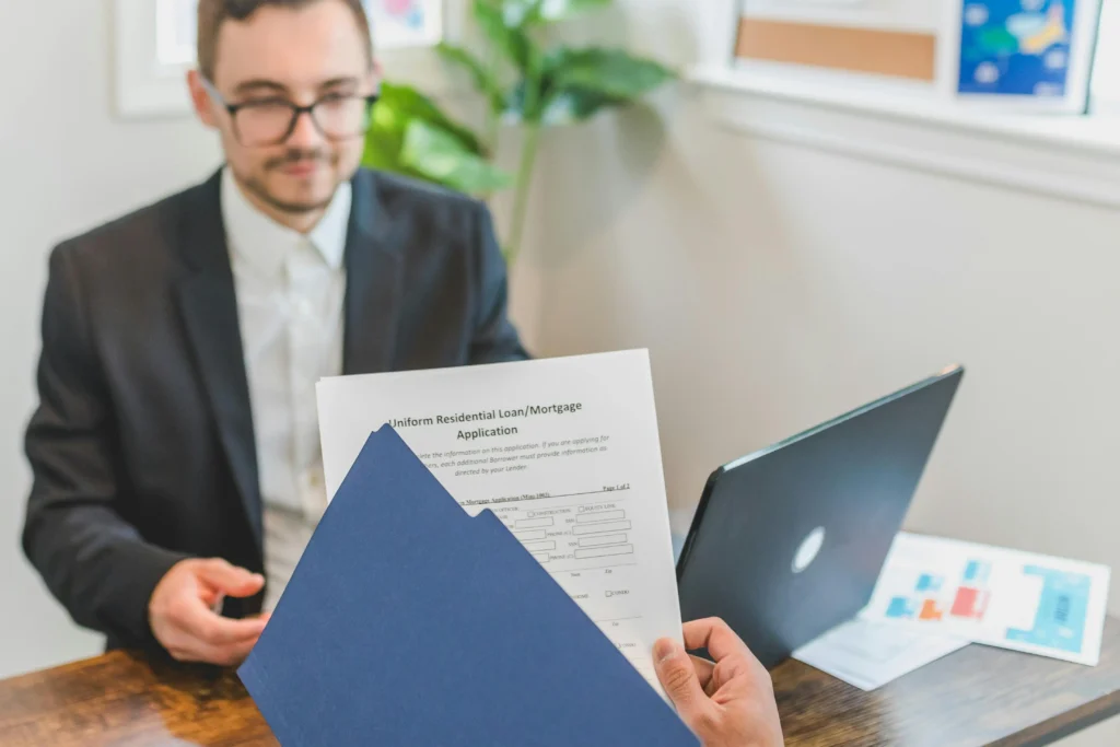A person in a suit holds a loan application, seated at a desk with a laptop and documents, in a well-lit office setting.